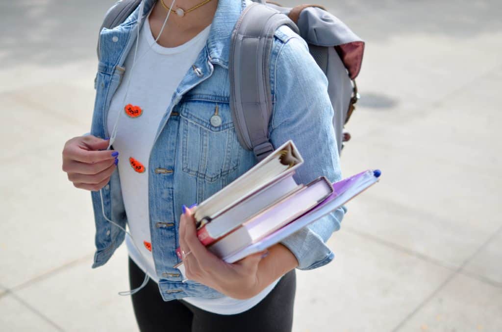 A student wearing a backpack, listening to music, and carrying a stack of books. 